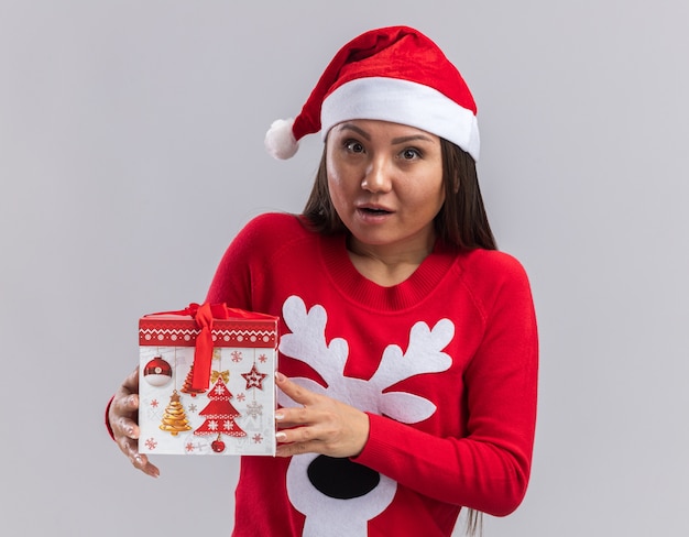 Preocupada joven asiática vistiendo gorro de navidad con un suéter con caja de regalo aislado sobre fondo blanco.