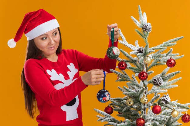 Preocupada joven asiática vistiendo gorro de navidad con suéter decorar el árbol de navidad aislado en la pared naranja