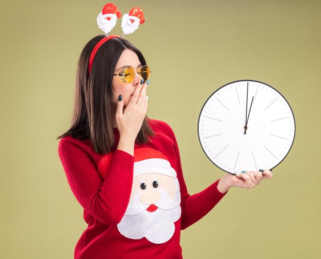 Preocupada joven bastante caucásica vistiendo suéter de santa claus y diadema con gafas sosteniendo y mirando el reloj manteniendo la mano en la boca aislada sobre fondo verde oliva