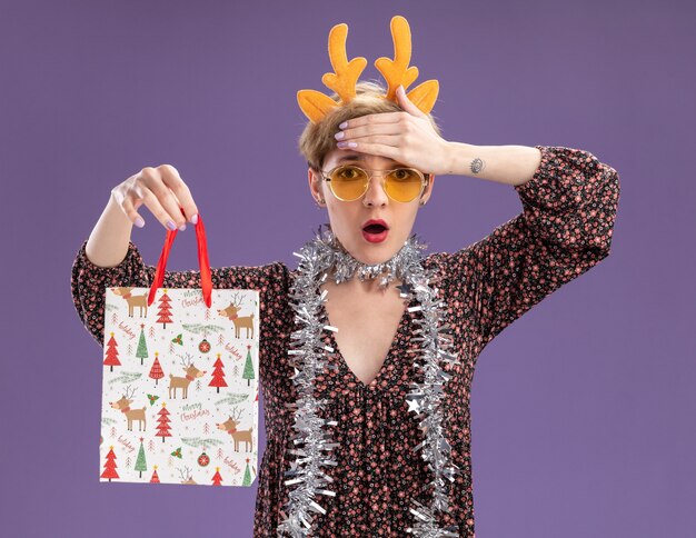 Preocupada joven bonita con diadema de cuernos de reno y guirnalda de oropel alrededor del cuello con gafas sosteniendo una bolsa de regalo de Navidad manteniendo la mano en la frente aislada en la pared púrpura