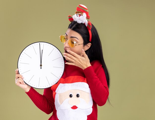 Preocupada joven caucásica con diadema de santa claus y suéter con gafas sosteniendo y mirando el reloj manteniendo la mano cerca de la boca aislada en la pared verde oliva con espacio de copia