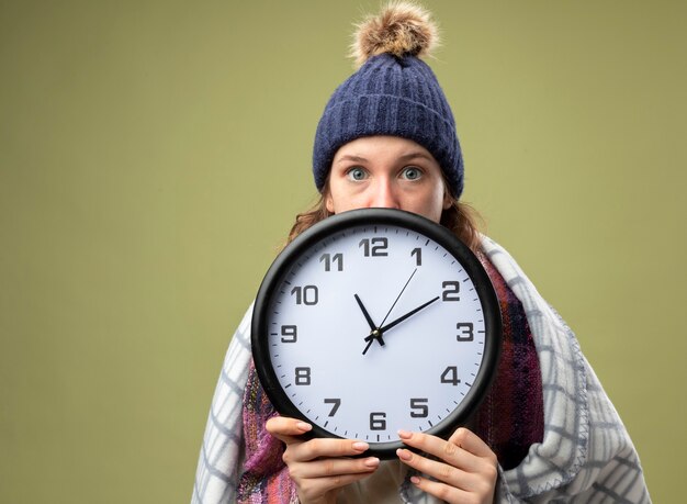 Preocupada joven enferma vestida con túnica blanca y gorro de invierno con la cara cubierta de bufanda con reloj de pared envuelto en cuadros aislados en verde oliva