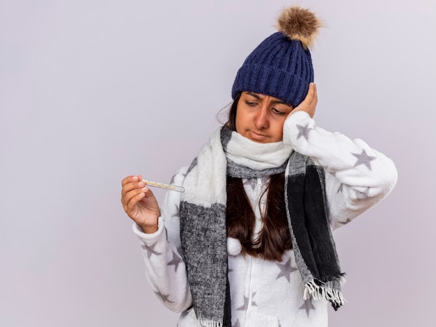 Preocupada joven enferma vistiendo gorro de invierno con bufanda sosteniendo y mirando el termómetro y poniendo la mano en la cabeza aislada sobre fondo blanco.