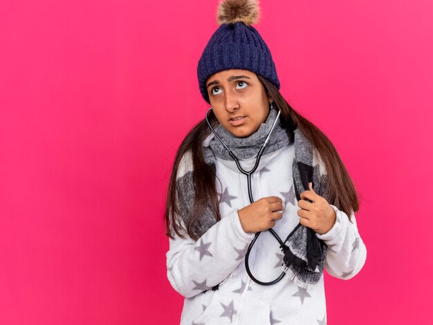 Preocupada joven enferma vistiendo gorro de invierno con bufanda vistiendo y escuchando sus propios latidos con estetoscopio aislado en rosa