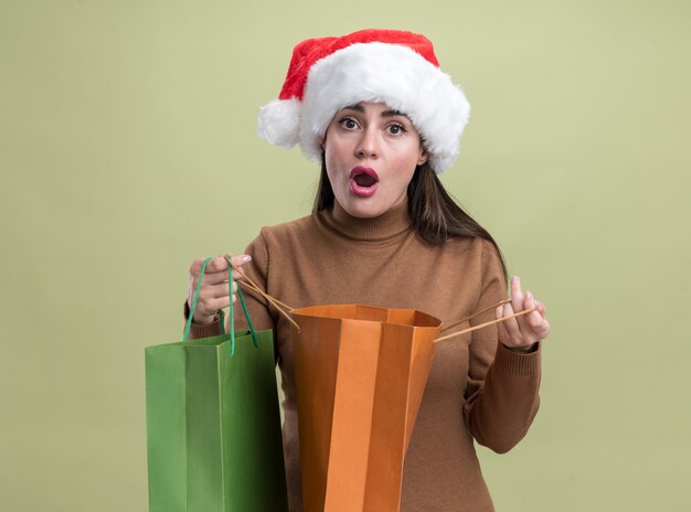 Foto gratuita preocupada joven hermosa niña con sombrero de navidad con bolsa de regalo aislado sobre fondo verde oliva