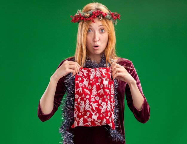 Preocupada joven hermosa vestida de rojo con corona y guirnalda en el cuello sosteniendo una bolsa de Navidad aislada en la pared verde