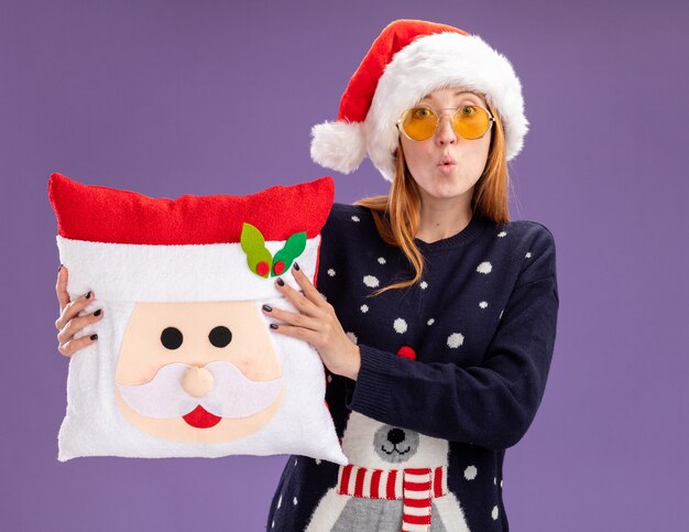 Preocupada joven hermosa vestida con suéter de Navidad y sombrero con gafas sosteniendo almohada de Navidad aislada en la pared púrpura