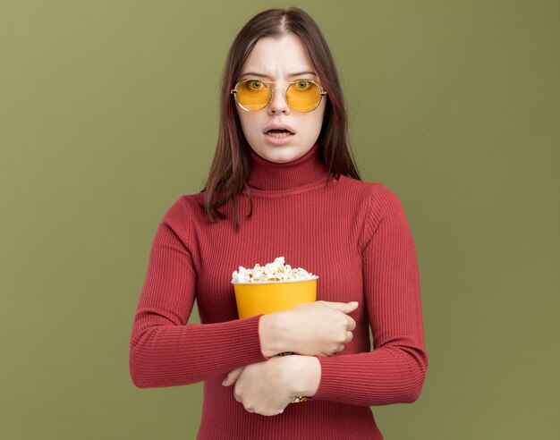 Preocupada joven mujer bonita con gafas de sol abrazando el cubo de palomitas de maíz