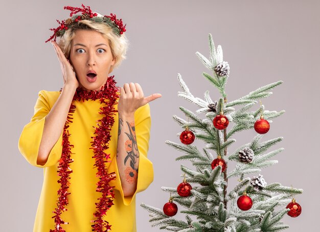Preocupada joven mujer rubia con corona de Navidad y guirnalda de oropel alrededor del cuello de pie cerca del árbol de Navidad decorado mirando manteniendo la mano cerca de la cara apuntando al lado aislado en la pared blanca