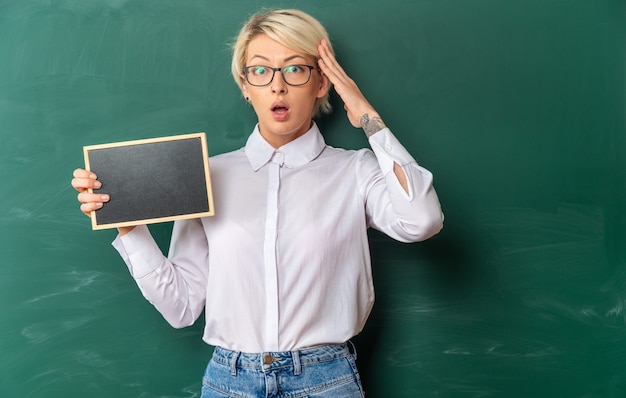 Preocupada joven profesora rubia con gafas en el aula de pie frente a la pizarra mostrando mini pizarra manteniendo la mano en la cabeza mirando al frente con espacio de copia