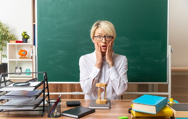 Preocupada joven profesora rubia con gafas sentado en un escritorio con herramientas escolares en el aula manteniendo las manos en la cara