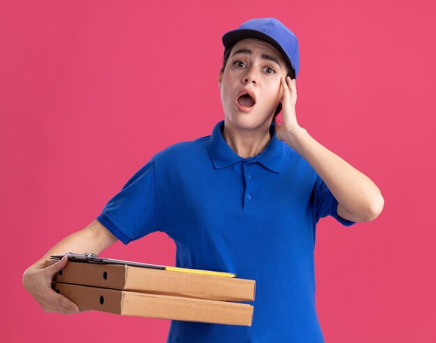 Preocupada joven repartidora en uniforme y gorra sosteniendo paquetes de pizza con portapapeles tocando la cara