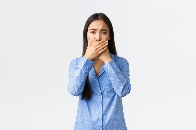 Preocupada linda chica asiática en pijama azul frunciendo el ceño y se ve preocupada mientras cierra la boca con las manos, cubre los labios, reacciona a noticias horribles, de pie, molesto fondo blanco.