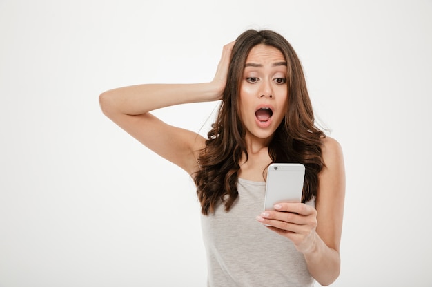 Preocupada mujer morena sorprendida mirando el teléfono inteligente mientras sostiene la cabeza sobre gris