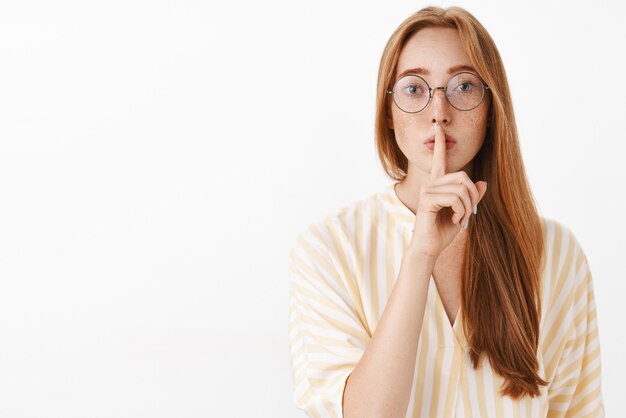 Preocupada mujer pelirroja tímida e insegura con pecas en gafas y elegante blusa de verano que pide no decirle a nadie diciendo shh con gesto de silencio sosteniendo el dedo índice sobre la boca