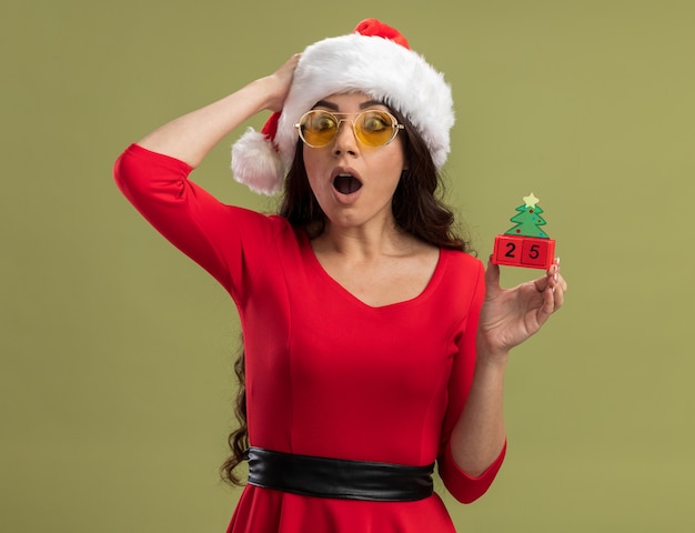 Preocupada niña bonita con gorro de Papá Noel y gafas sosteniendo el juguete del árbol de Navidad con fecha manteniendo la mano en la cabeza mirando al lado aislado en la pared verde oliva