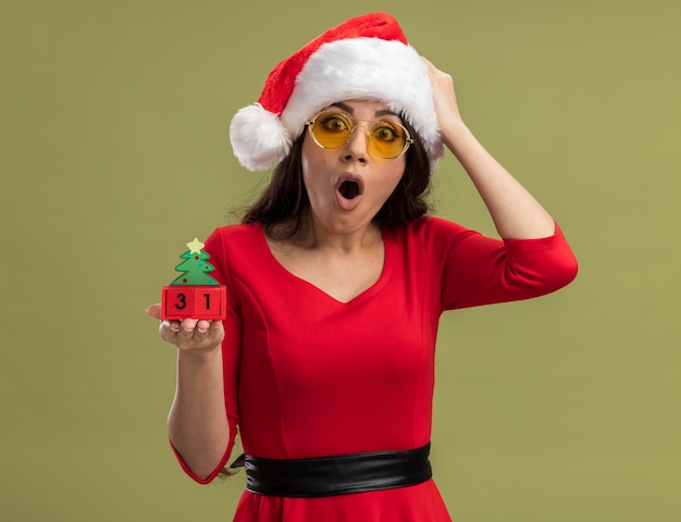 Foto gratuita preocupada niña bonita vistiendo gorro de papá noel y gafas sosteniendo el juguete del árbol de navidad con fecha poniendo la mano en la cabeza mirando a la cámara aislada sobre fondo verde oliva