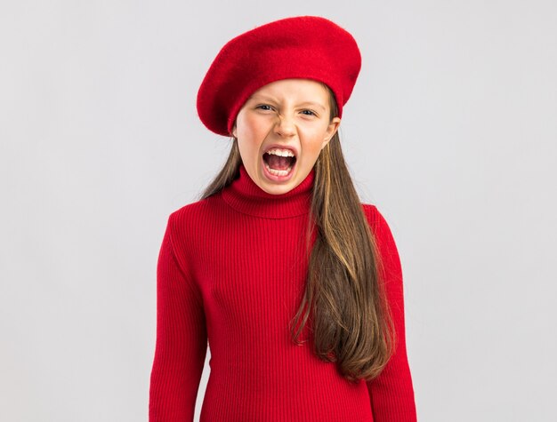 Preocupada niña rubia vistiendo boina roja mirando al frente y gritar aislado en la pared blanca con espacio de copia