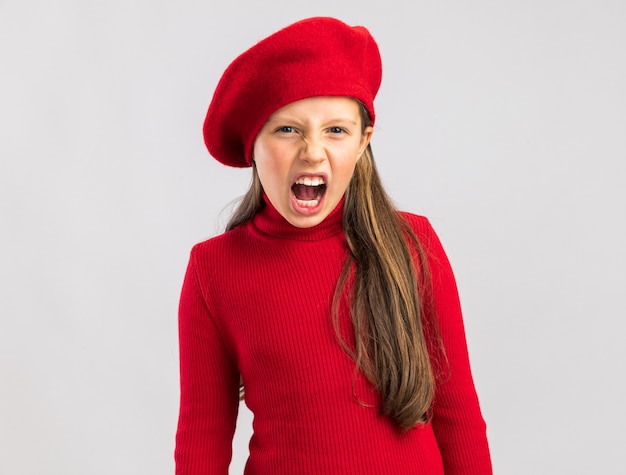 Preocupada niña rubia vistiendo boina roja mirando al frente y gritar aislado en la pared blanca con espacio de copia