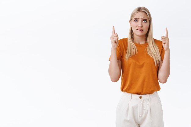 Preocupada, preocupada joven linda mujer rubia insegura con camiseta naranja, pantalones, mordiéndose el labio con ansiedad, frunciendo el ceño y mirando hacia arriba, señalando el anuncio superior, sintiéndose nervioso, fondo blanco