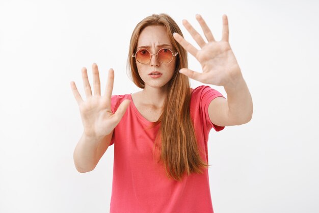 Preocupada y preocupada joven pelirroja con gafas de sol y camiseta rosa que comienza a ponerse nerviosa abriendo la boca frunciendo el ceño levantando las palmas cerca del pecho como si estuviera atrapada en una caja, refrenando a un hombre peligroso