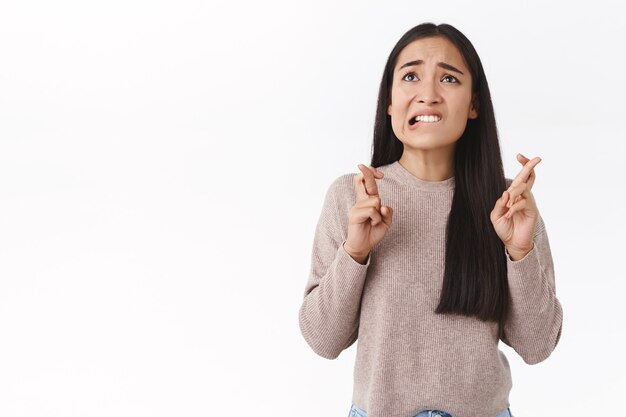 Preocupada, preocupada linda chica asiática con blusa beige, tiene preocupaciones, cruza los dedos, buena suerte, muerde el labio nerviosamente mira el gráfico, espera resultados importantes, ansiosamente espero que el deseo se haga realidad