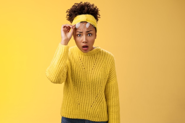 Preocupada sorprendida sorprendida estudiante afroamericana sin palabras caída de la mandíbula gafas de despegue abrir los ojos sorprendido ha sido abandonado mirar algo increíble, de pie cuestionado frustrado, fondo amarillo.