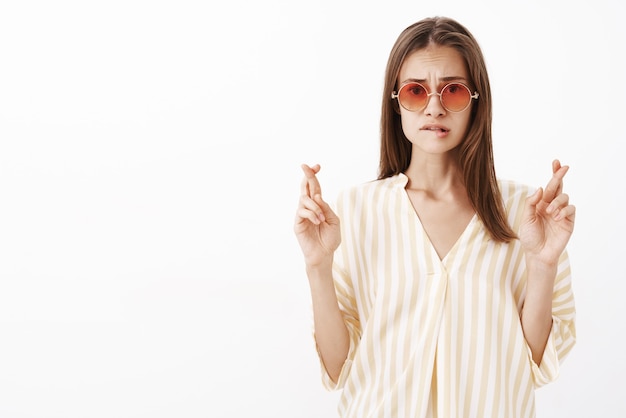 Preocupada, triste y guapa mujer elegante en una blusa de rayas amarillas de moda y gafas de sol, mordiéndose el labio frunciendo el ceño y cruzando los dedos para la buena suerte mientras pide deseos y espera con suerte el resultado