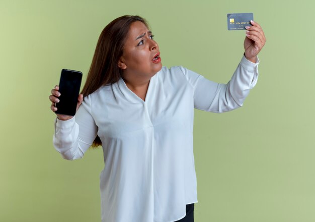 Preocupado casual mujer de mediana edad caucásica sosteniendo el teléfono y mirando la tarjeta de crédito en su mano