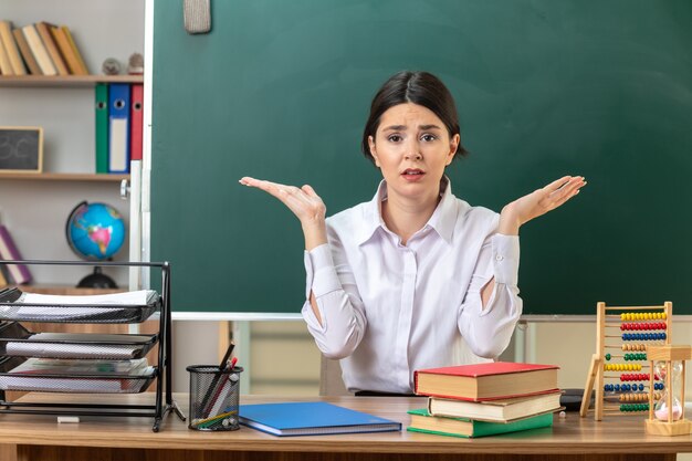 Preocupado extendiendo las manos joven maestra sentada a la mesa con herramientas escolares en el aula