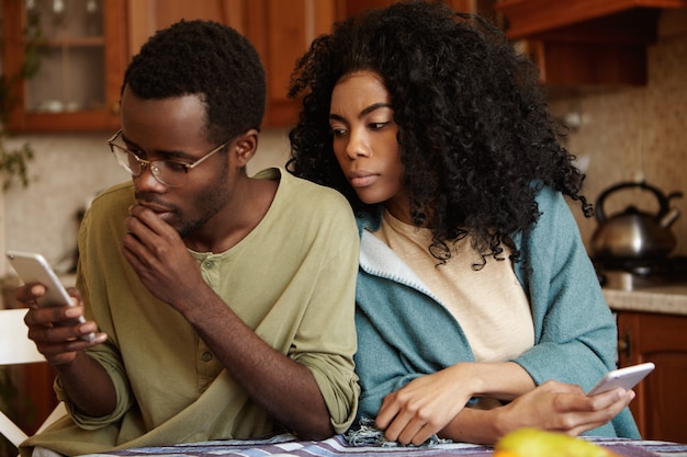 Preocupado joven afroamericano con gafas escribiendo sms en el teléfono inteligente sumido en sus pensamientos sin darse cuenta de que su novia está espiando, mirando por encima del hombro, tratando de leer lo que está enviando mensajes de texto