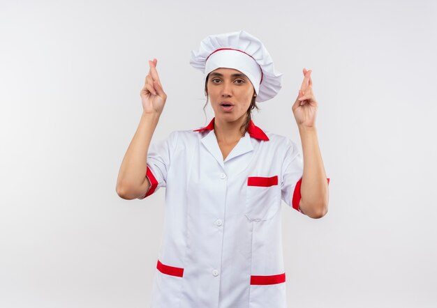 Preocupado joven cocinera vistiendo uniforme de chef cruzando los dedos sobre la pared blanca aislada con espacio de copia