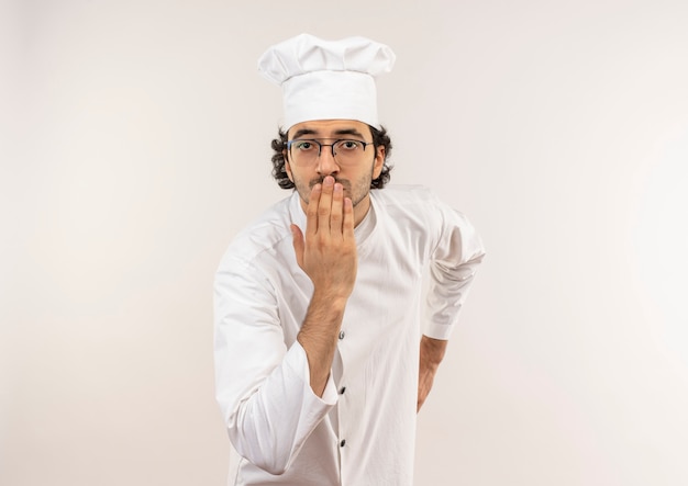 Preocupado joven cocinero vistiendo uniforme de chef y gafas tapadas la boca con la mano