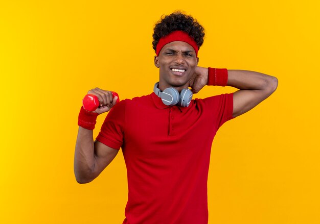 Preocupado joven deportivo afroamericano con diadema y muñequera con auriculares en el cuello sosteniendo mancuernas y poniendo la mano detrás de la cabeza aislada en la pared amarilla