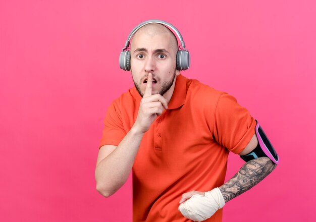 Preocupado joven deportivo con vendaje de muñeca usando audífonos con brazalete de teléfono mostrando gesto de silencio aislado en la pared rosa