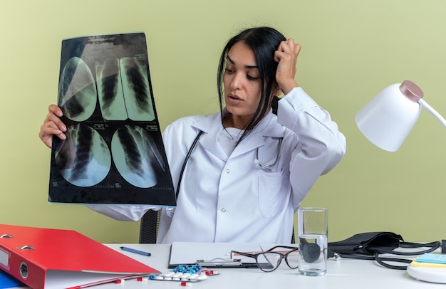 Preocupado joven doctora vistiendo bata médica con estetoscopio se sienta en el escritorio con herramientas médicas sosteniendo rayos x poniendo la mano en la cabeza aislada en la pared verde oliva