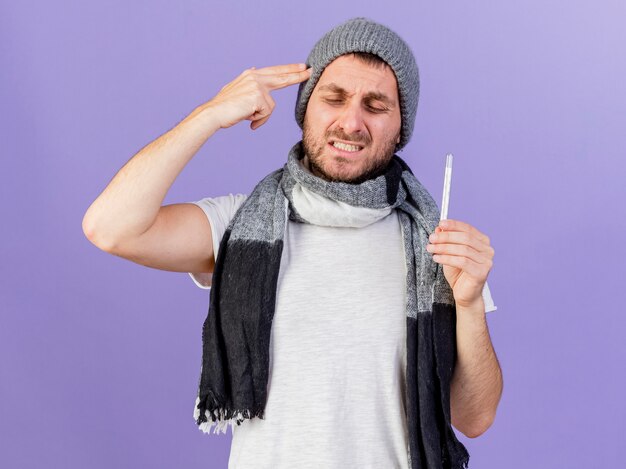 Preocupado joven enfermo vistiendo gorro de invierno con bufanda sosteniendo el termómetro y poniendo los dedos en la sien aislado en púrpura