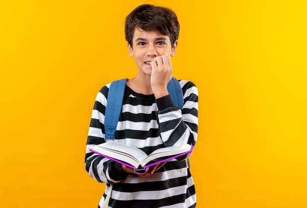 Preocupado joven escolar con mochila sosteniendo libro muerde las uñas aislado en la pared naranja