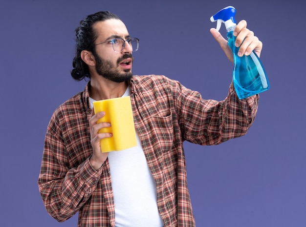 Preocupado joven guapo chico de limpieza con camiseta sosteniendo una esponja mirando la botella de spray en la mano aislada en la pared azul