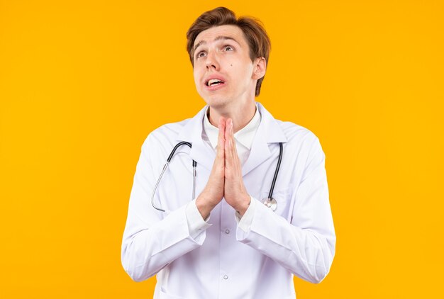 Preocupado joven médico vistiendo bata médica con estetoscopio mostrando gesto de oración aislado en la pared naranja