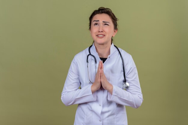 Preocupado joven médico vistiendo bata médica con estetoscopio muestra gesto de oración en la pared verde