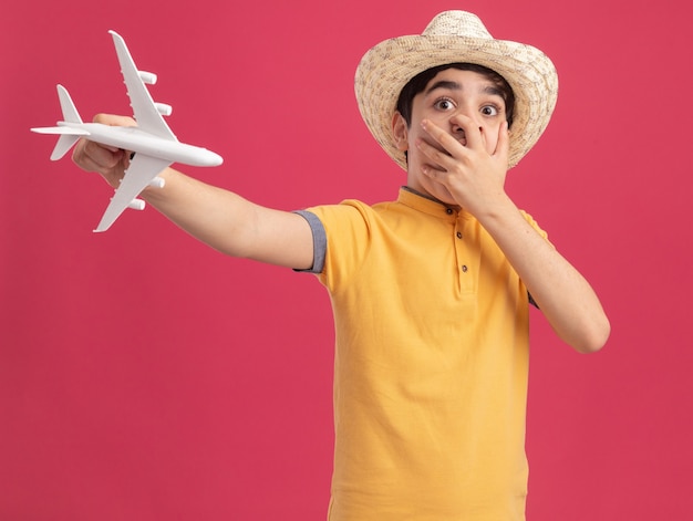 Foto gratuita preocupado joven muchacho caucásico con sombrero de playa estirando el modelo de avión mirando recto manteniendo la mano en la boca