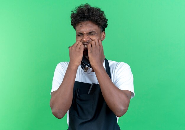Preocupado joven peluquero afroamericano vistiendo uniforme muerde las uñas aislado sobre fondo verde