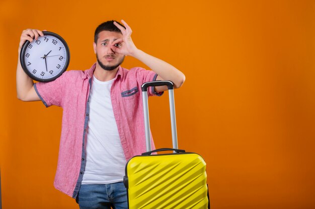 Preocupado joven viajero guapo chico con reloj de pie con la maleta haciendo un signo bien y mirando a través de este signo con expresión triste en la cara sobre fondo naranja