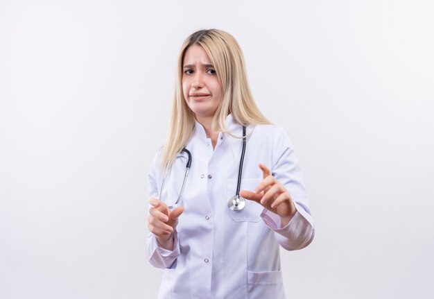 Preocupado médico joven rubia con estetoscopio y bata médica que muestra el tamaño con ambas manos en la pared blanca aislada