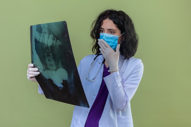 Foto gratuita preocupado médico joven vistiendo bata blanca con estetoscopio en máscara protectora médica mirando nervioso en la radiografía de los pulmones de pie en verde aislado