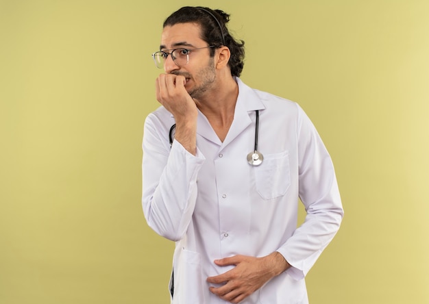 Preocupado médico varón joven con gafas ópticas vistiendo túnica blanca con estetoscopio muerde las uñas