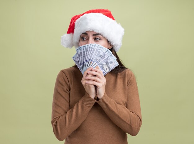 Preocupado mirando al lado hermosa joven con sombrero de navidad rostro cubierto con dinero en efectivo aislado en la pared verde oliva