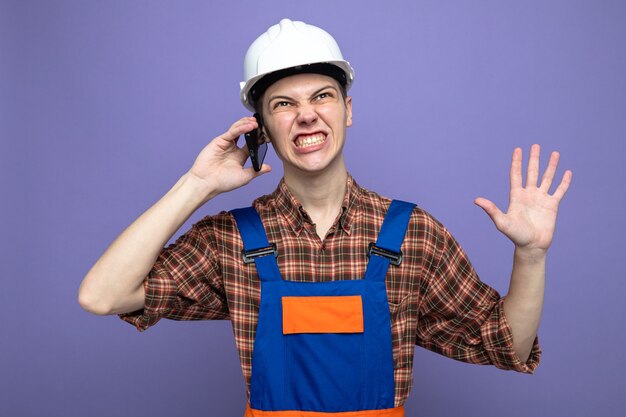 Preocupado mostrando cinco jóvenes constructores masculinos vistiendo uniforme habla por teléfono