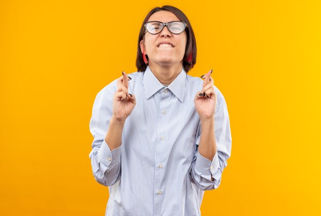 Preocupado por los ojos cerrados joven hermosa con gafas cruzando los dedos aislados en la pared naranja